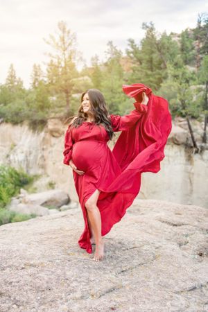 Woman in red dress twirls on rocky outcrop with dramatic mountain scenery behind her.
