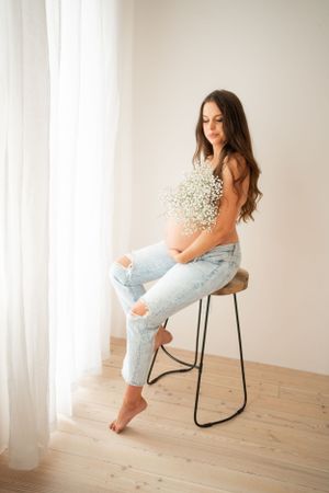 A woman in ripped jeans sitting on a stool by a window holding baby's breath flowers.