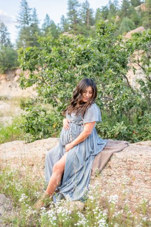 Woman in flowing blue dress poses gracefully against blooming bushes in a mountain setting.