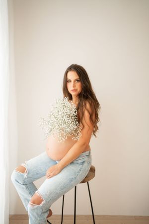 Woman with baby's breath flowers poses on stool in ripped jeans during maternity shoot.