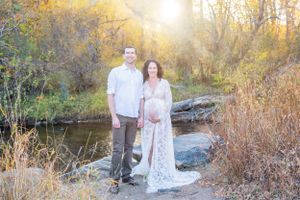 A maternity photoshoot by a stream featuring a white lace gown in autumn scenery.