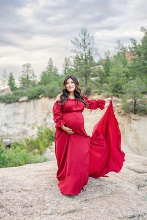 Expectant mother in red flowing gown poses dramatically on rocky cliff with forest backdrop.