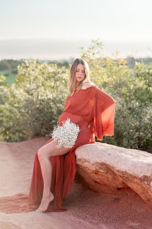A flowing red dress catches sunlight as someone poses with baby's breath flowers on red rocks at sunset.
