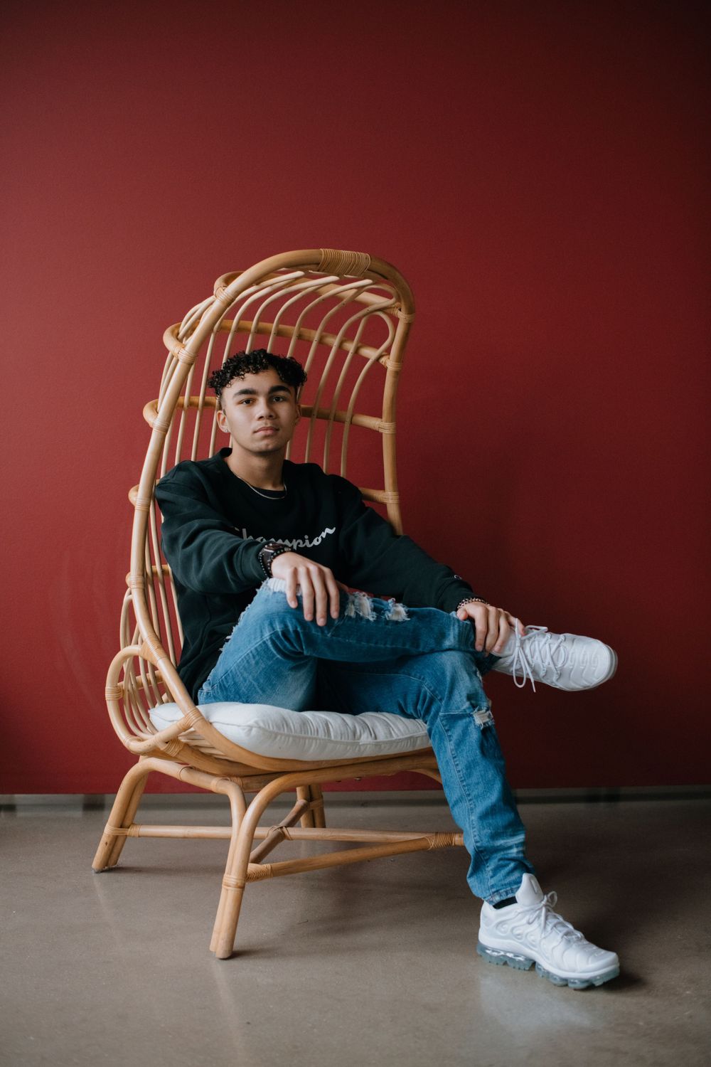 Stylish person in casual wear sitting on rattan chair against red wall.