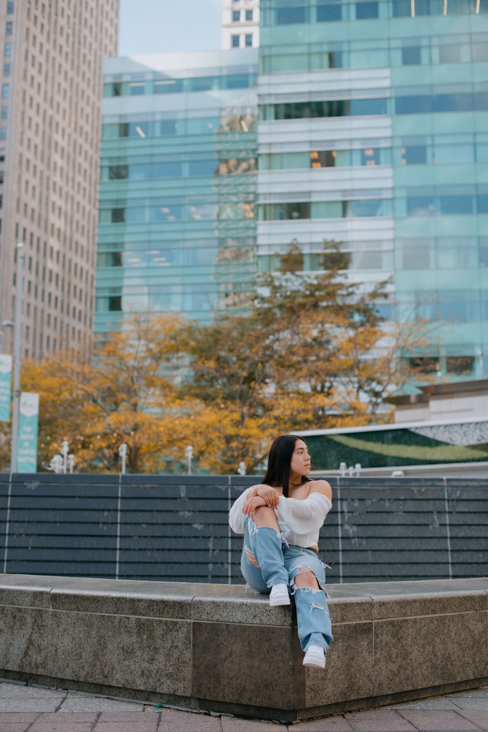 Relaxed urban portrait on concrete ledge with glass buildings and autumn trees in background.