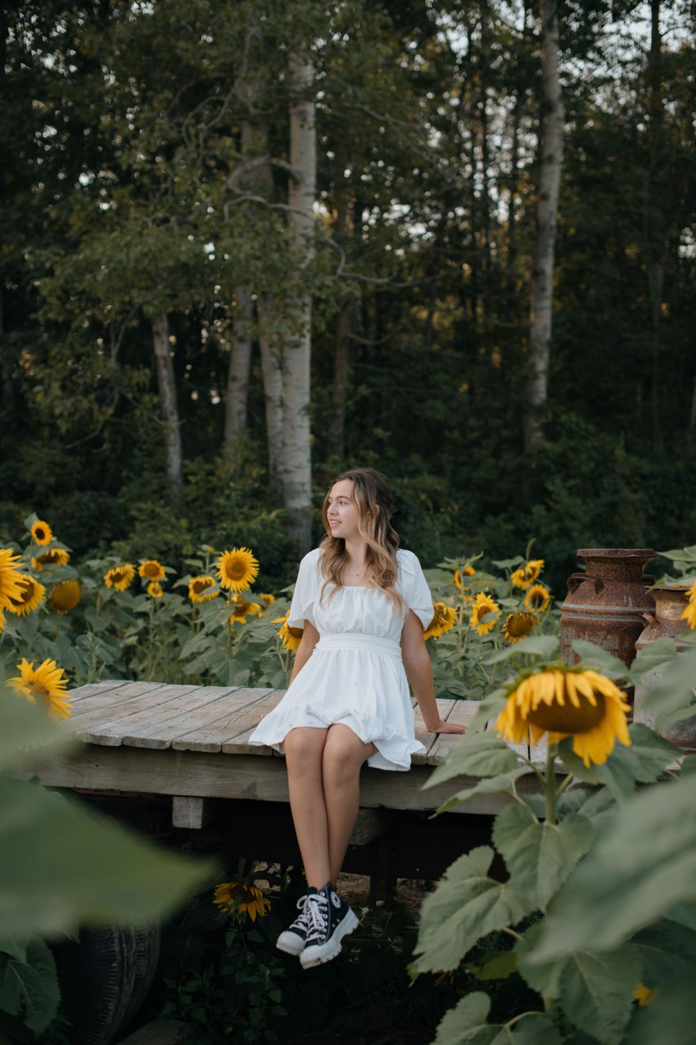 A dreamy garden scene with someone in white sitting on a wooden deck surrounded by sunflowers.