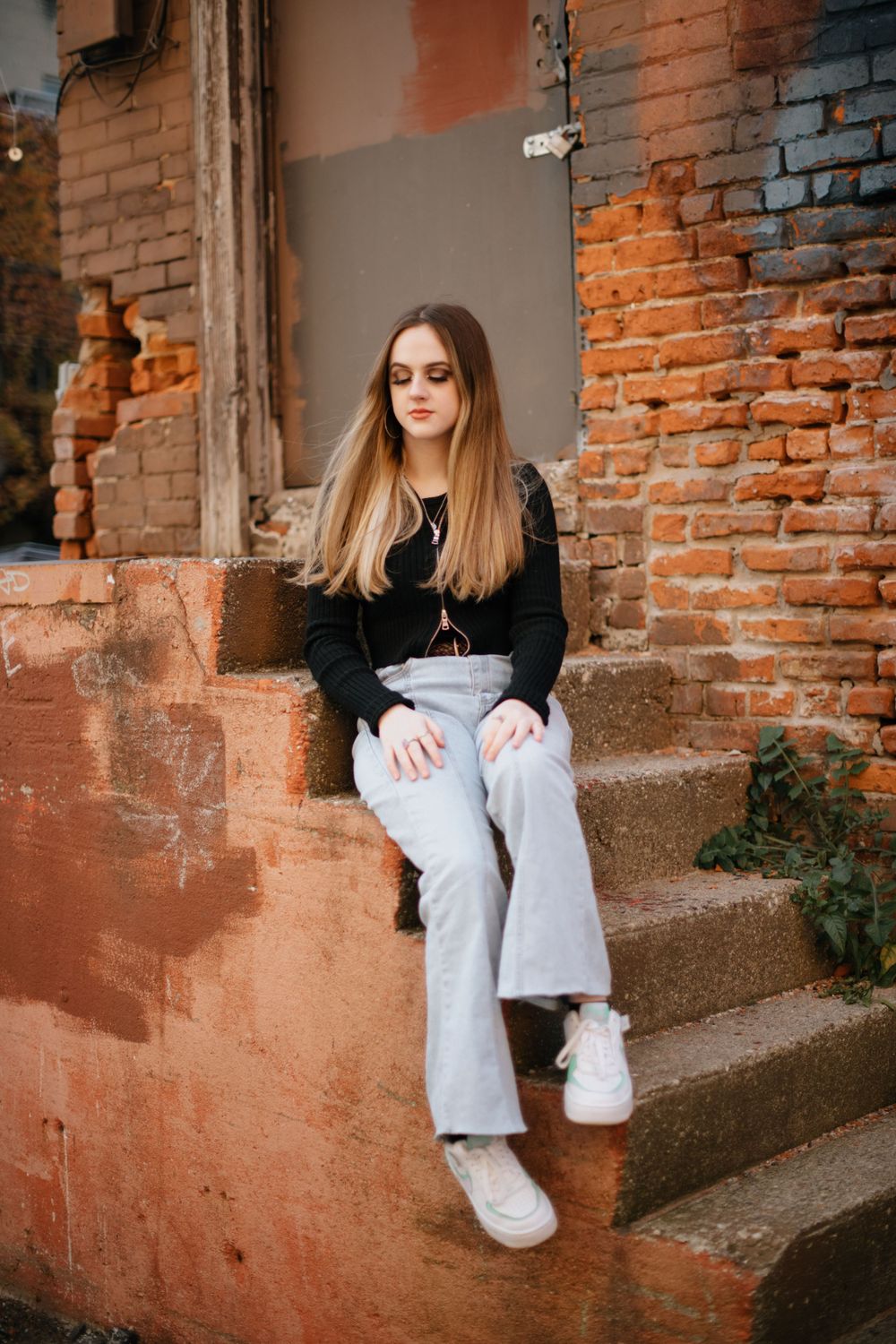 Casual portrait on brick steps showing person in black sweater and light jeans with white sneakers.