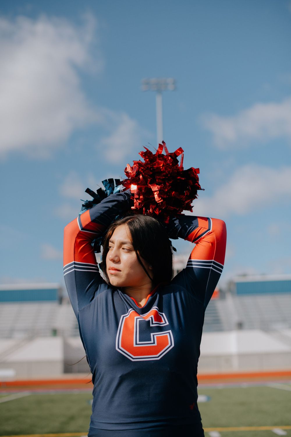 Cheerleader in navy uniform holding red pom poms overhead on sports field.