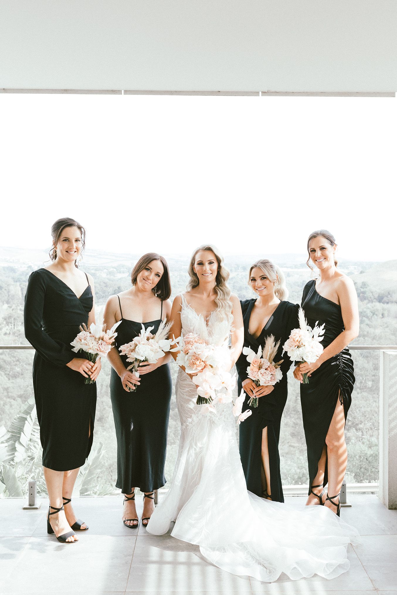 Elegant bridal party wearing black dresses and holding floral bouquets pose together on a balcony with mountain views.