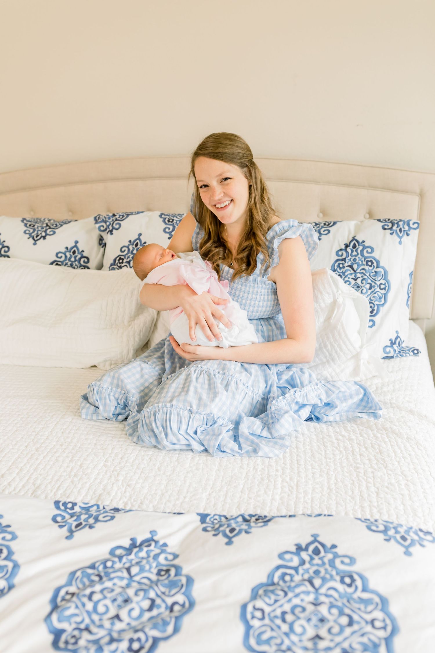 Mother in blue dress holds newborn baby while sitting on bed with patterned comforter.