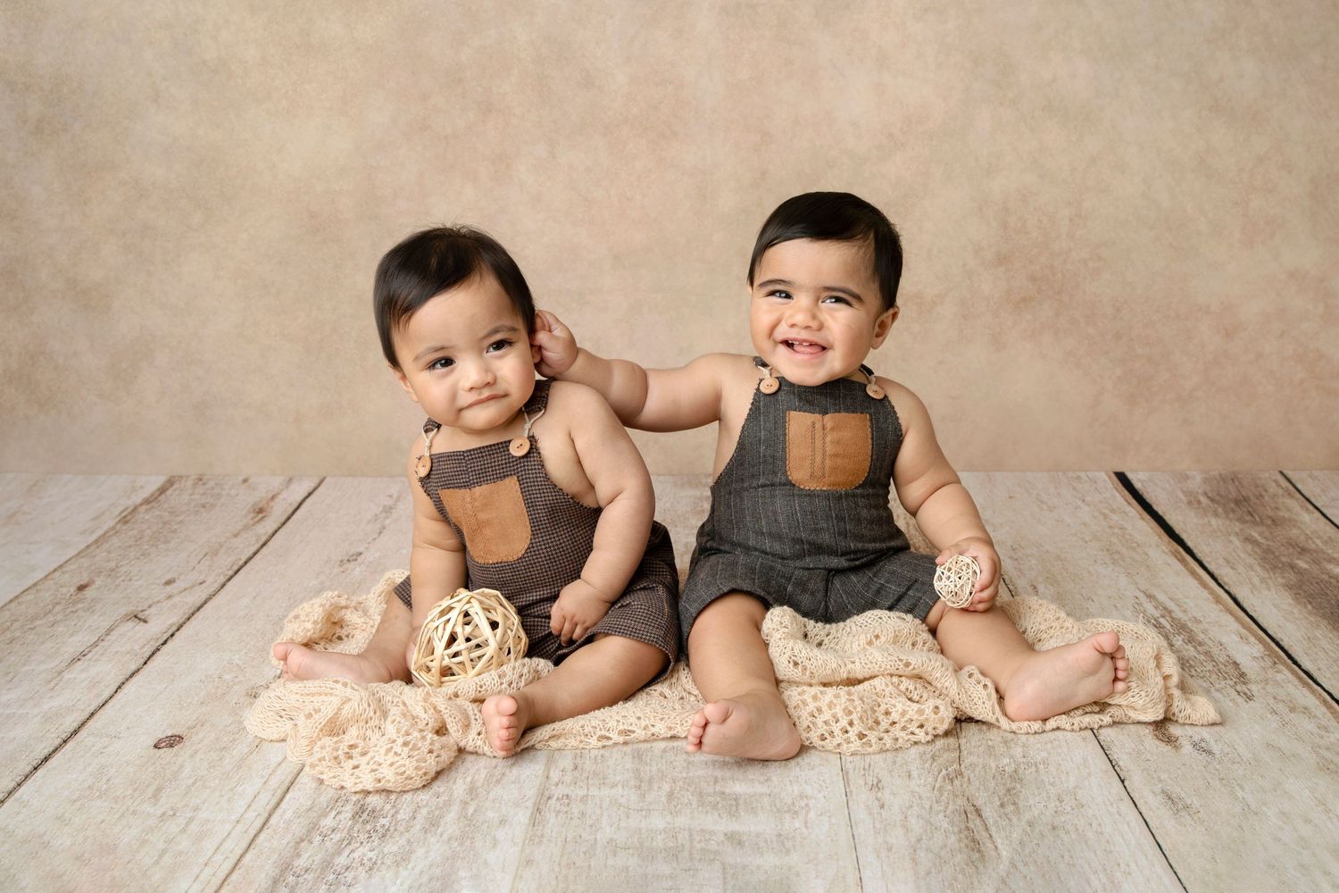 Two babies in matching brown overalls sitting on wooden floor for portrait.