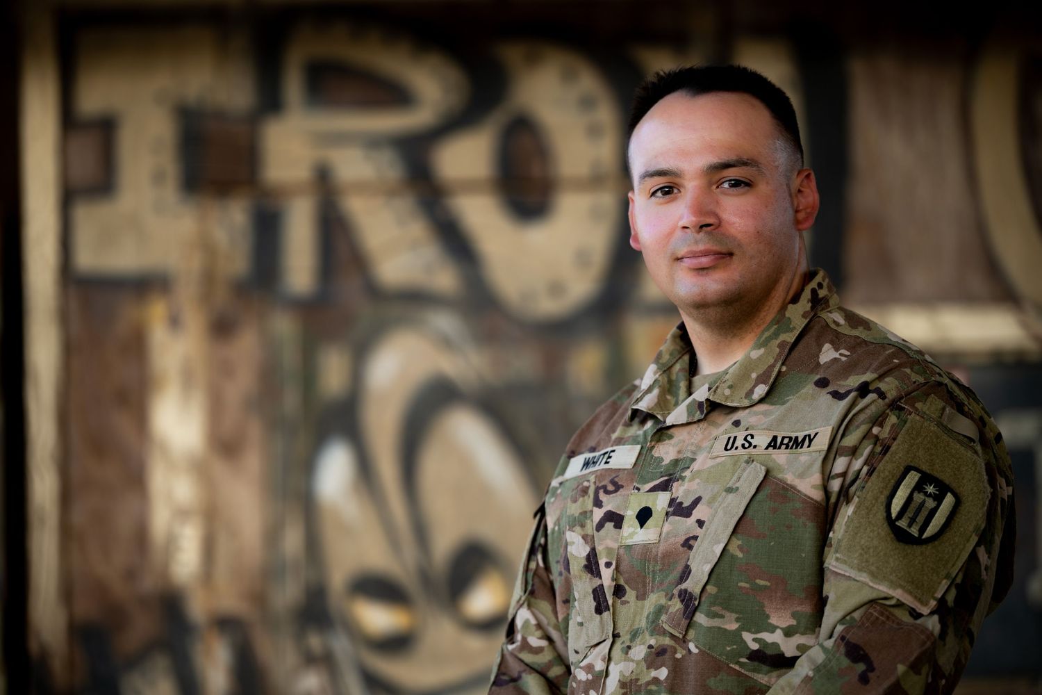 U.S. Army soldier in multicam uniform smiling in front of a graffiti-covered wall.