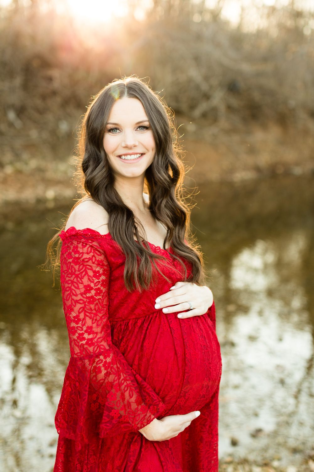 Maternity portrait in red lace dress against water and sunset backdrop.