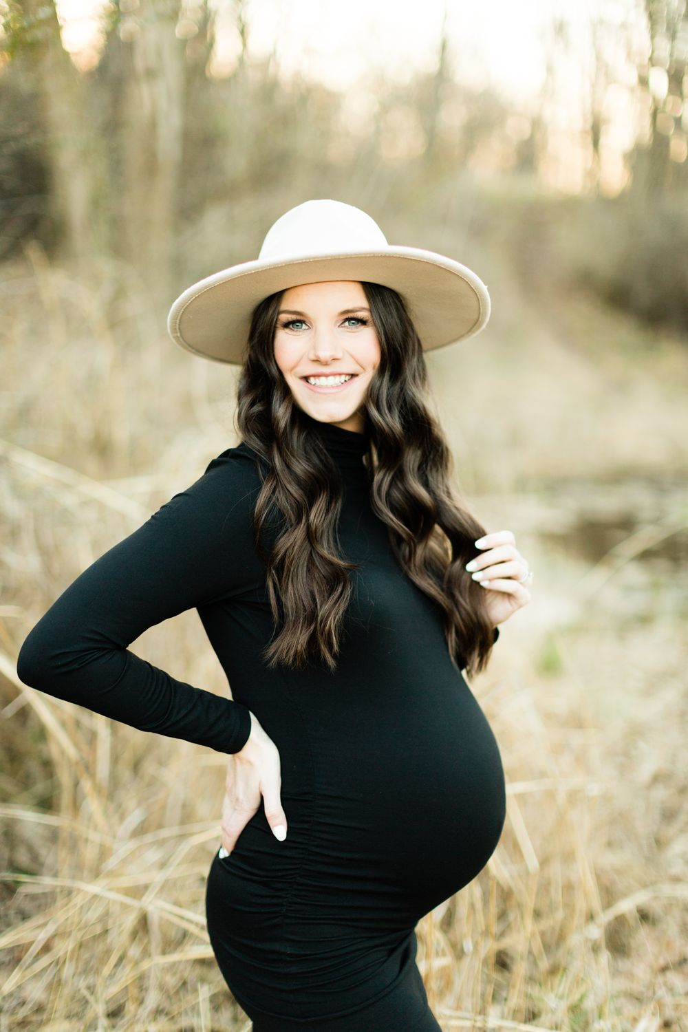 Maternity portrait of woman in black dress and wide brim hat posing in natural setting.