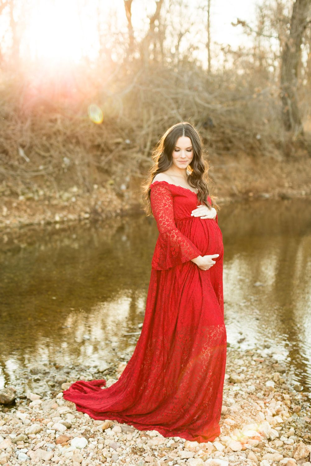 A stunning maternity photo in a flowing red lace dress against a reflective pond at golden hour.