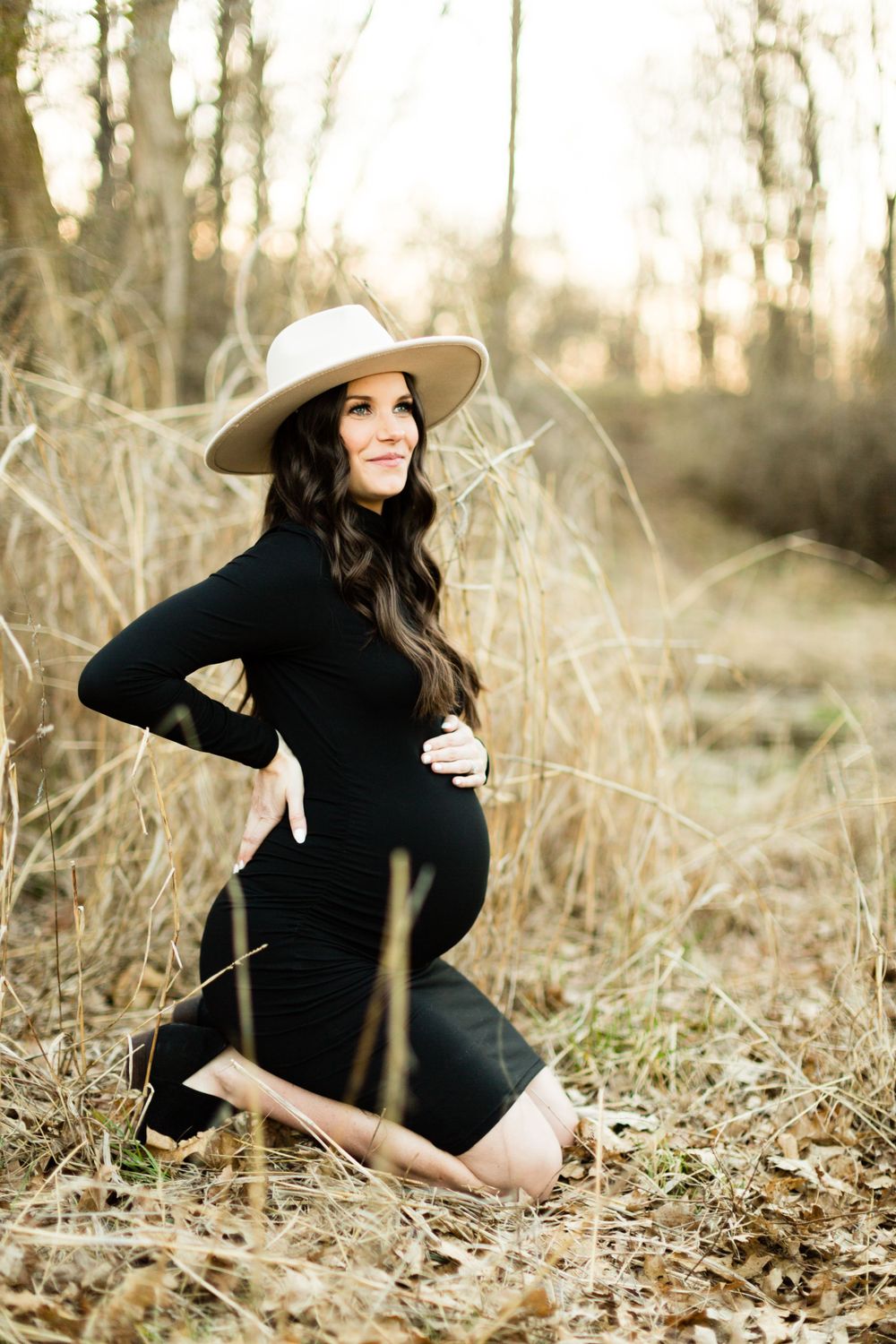 Elegant maternity portrait in black dress and hat among dried winter grass.