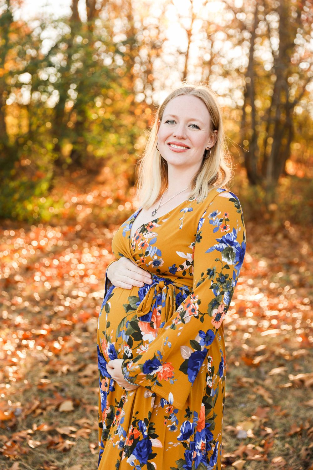 A woman in a yellow floral dress stands smiling in a forest filled with golden autumn leaves.