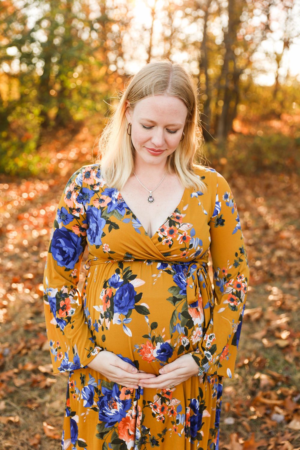 A pregnant woman in a mustard yellow floral dress poses in a sun-dapped autumn forest setting.