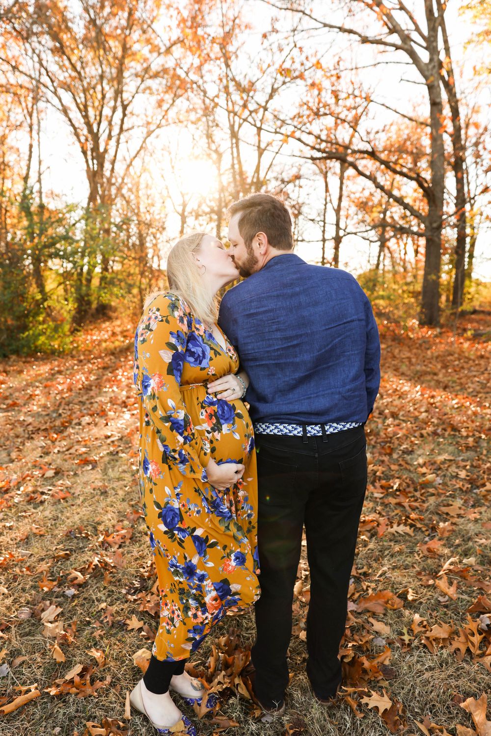 A couple embraces while standing among fallen autumn leaves in a wooded area at sunset.