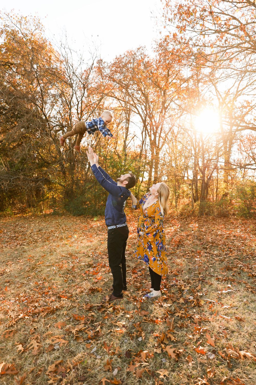 A couple plays with their child in a sun-lit autumn forest, lifting them joyfully in the air.