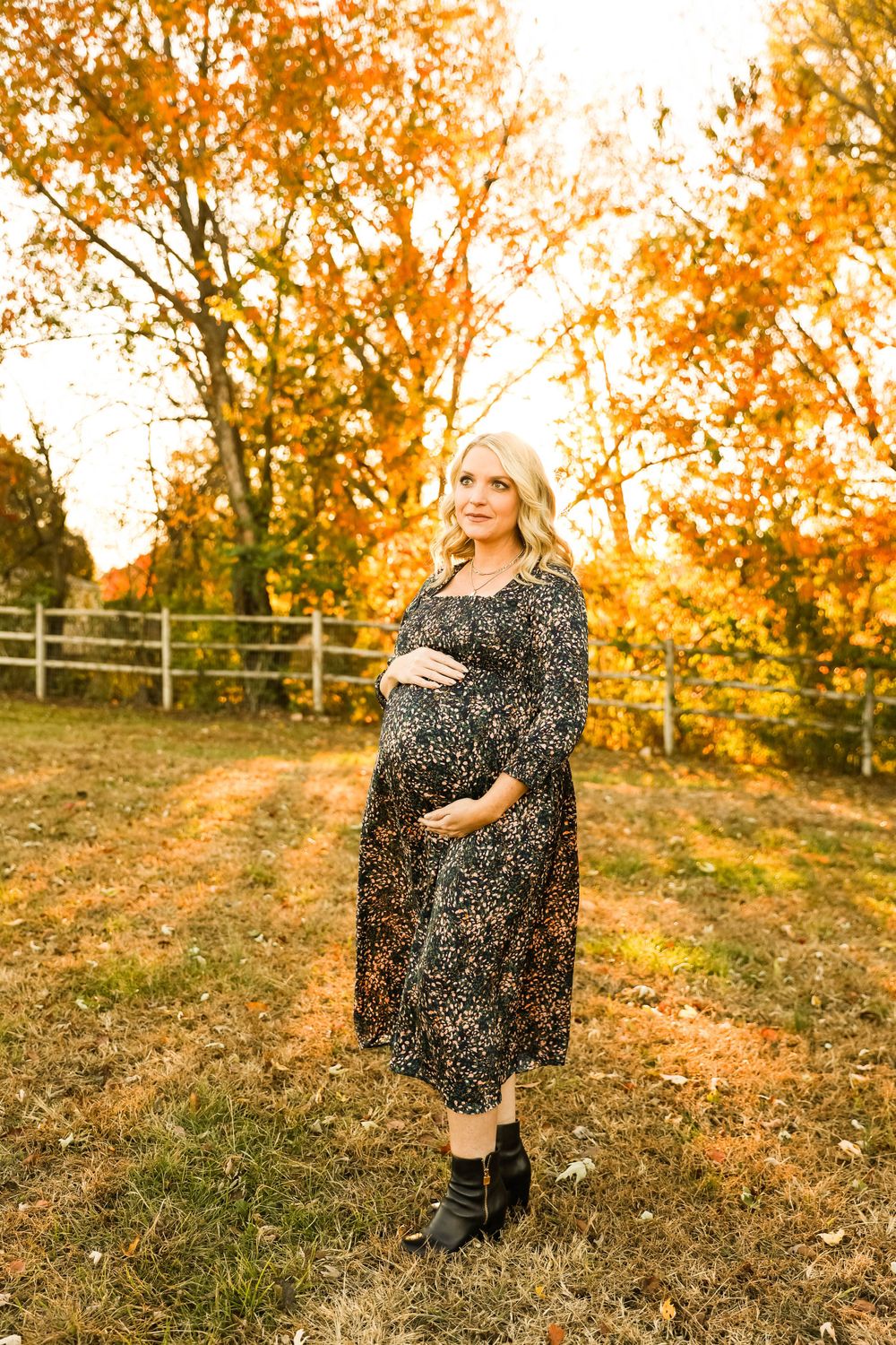Expectant mother in a floral dress poses in front of autumn trees and a rustic fence.