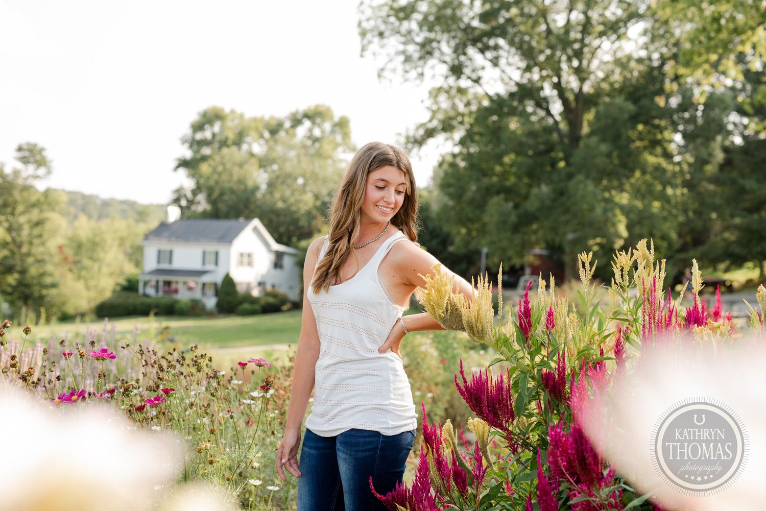 A person poses casually in a white tank top among vibrant red and pink flowers in a backyard garden setting.