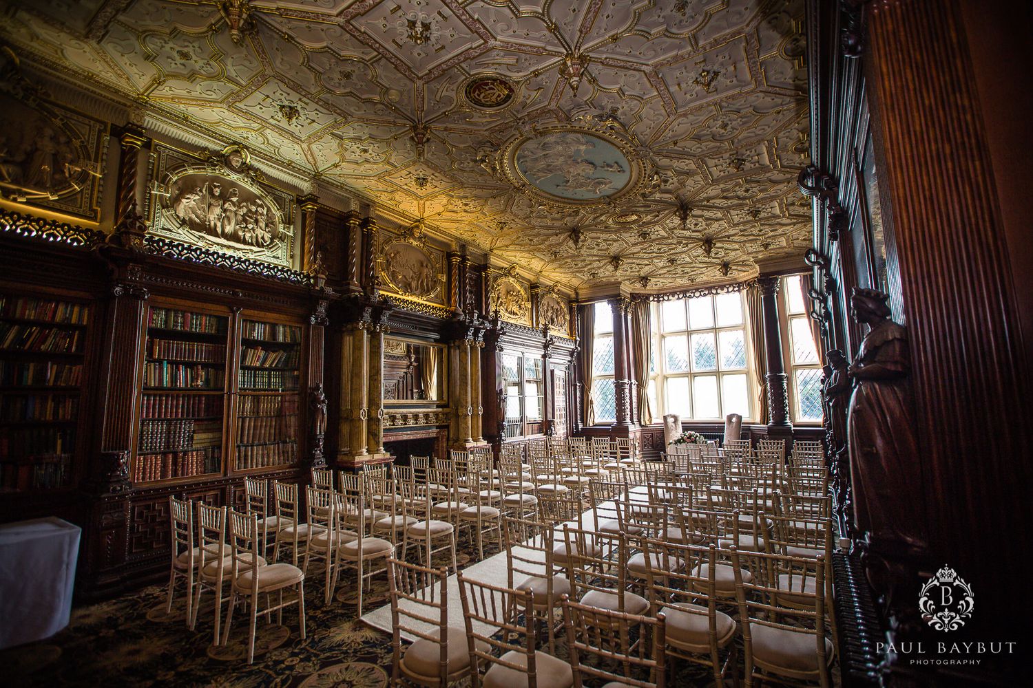 Ornate historic ballroom with decorative ceiling and rows of white chairs set up for an elegant ceremony.