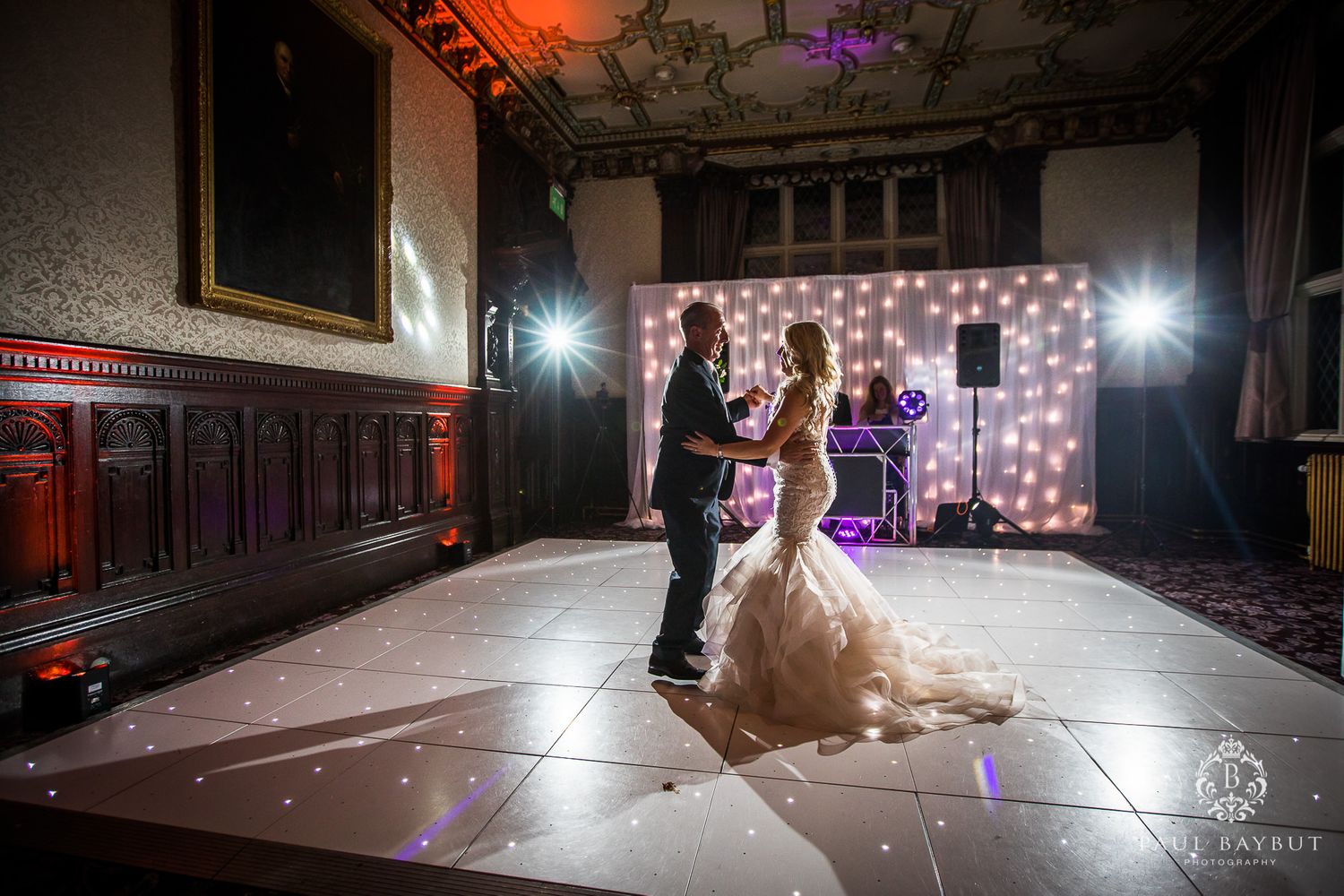 A wedding couple dances beneath spotlights with a twinkling light wall creating magical ambiance.