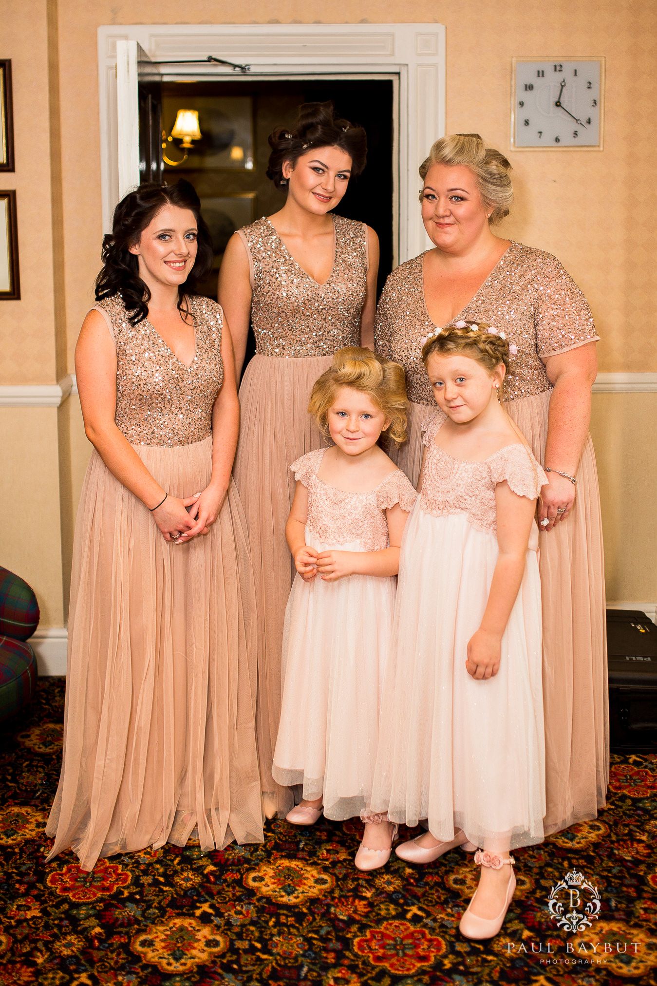Bridesmaids and flower girls pose together in matching blush pink dresses on a patterned carpet at a wedding venue.