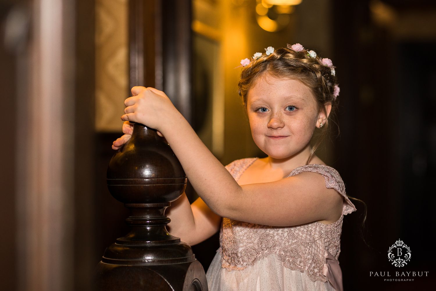 Young flower girl in a pink dress and floral crown poses with a decorative banister.