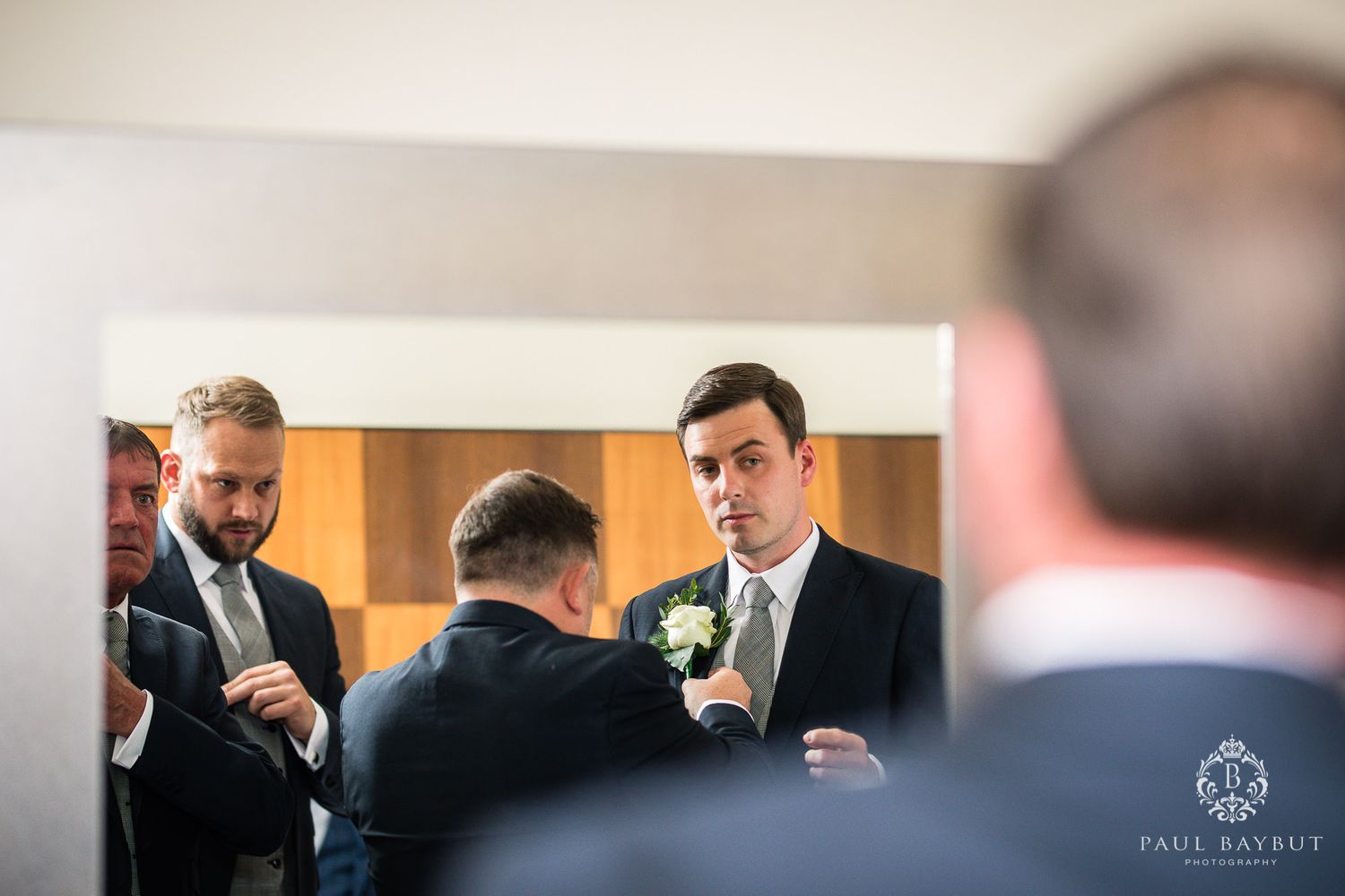Wedding preparation scene reflected in mirror shows groomsmen adjusting formal attire.