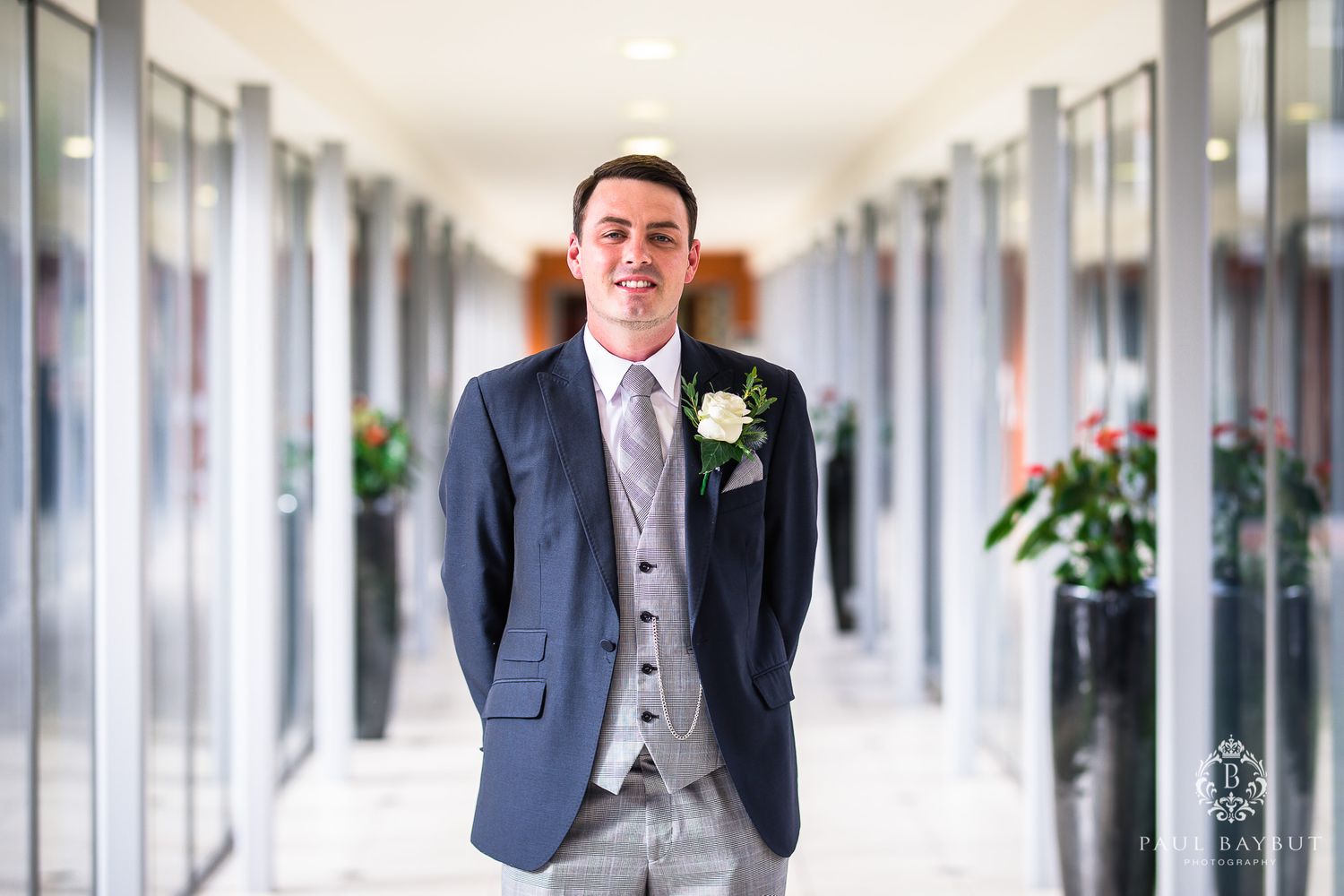 Groom in navy suit and grey waistcoat poses in bright hallway with boutonniere.