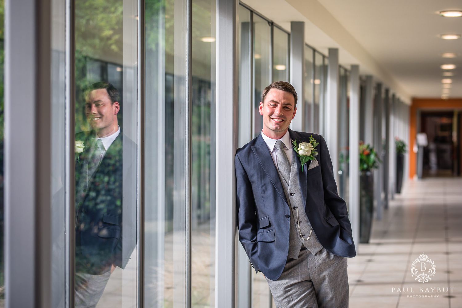 Groom in formal attire poses against glass windows in modern hallway with reflection.