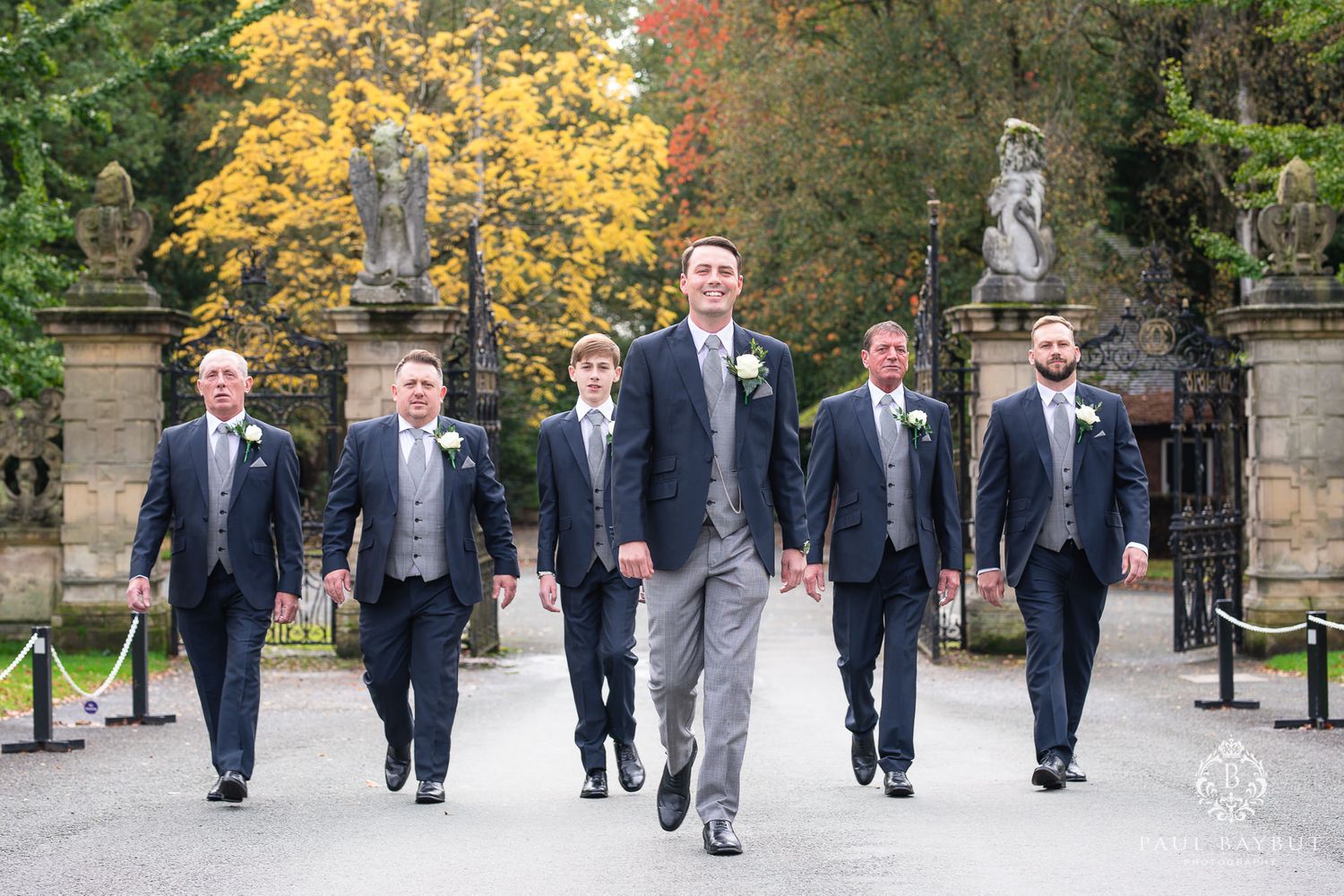 Groomsmen walk together on stone path with autumn foliage and statues in background.