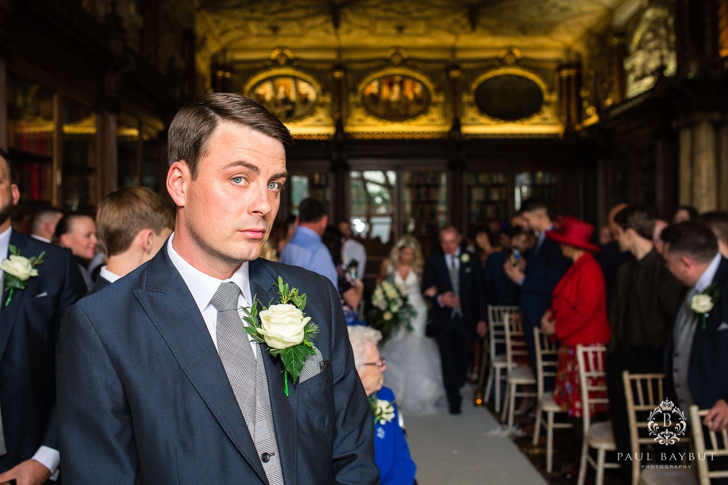 A groomsman in a navy suit with white boutonniere stands in an ornate library venue.
