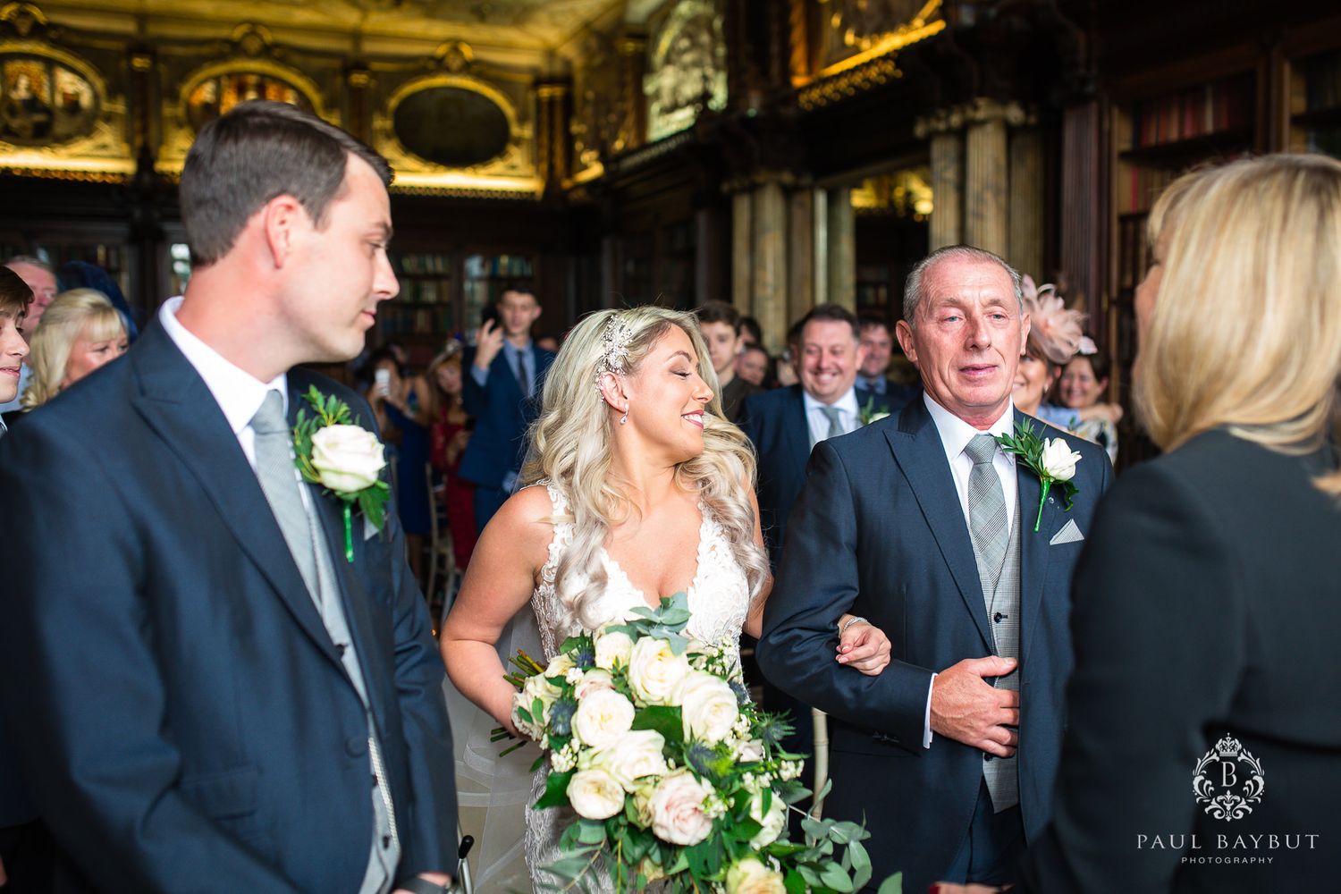 A wedding ceremony takes place in an elegant library with gold ornate ceiling and bookshelves.