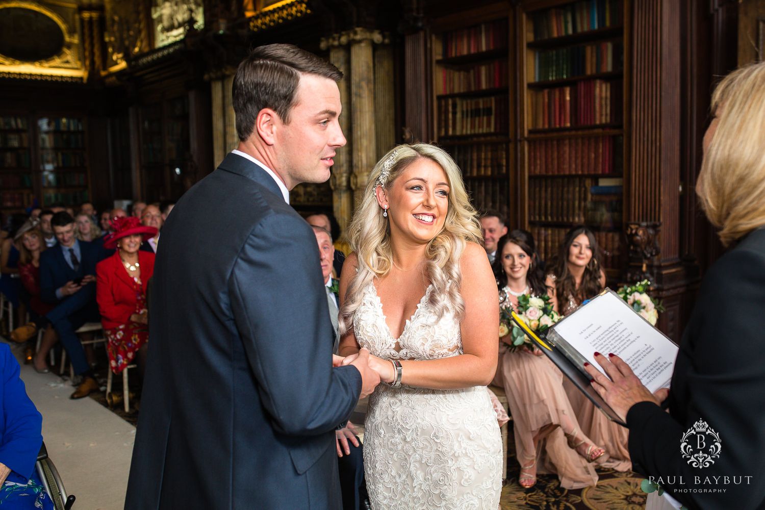 A couple exchanges vows in front of guests in a grand library setting.