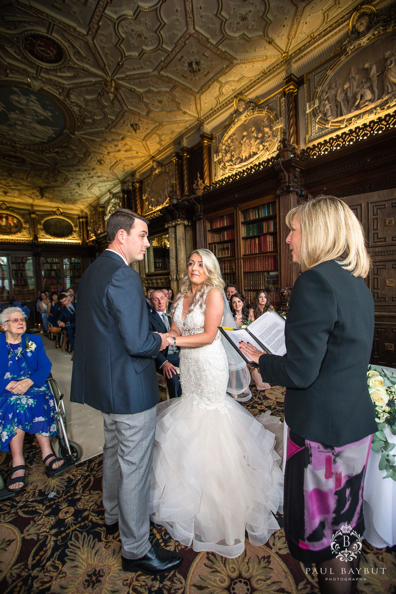 Wedding ceremony taking place in a grand historic library with ornate ceiling and bookshelves.