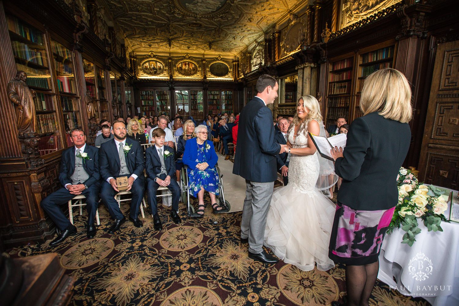 Wedding ceremony in a vintage library setting with guests seated on ornate carpeting.