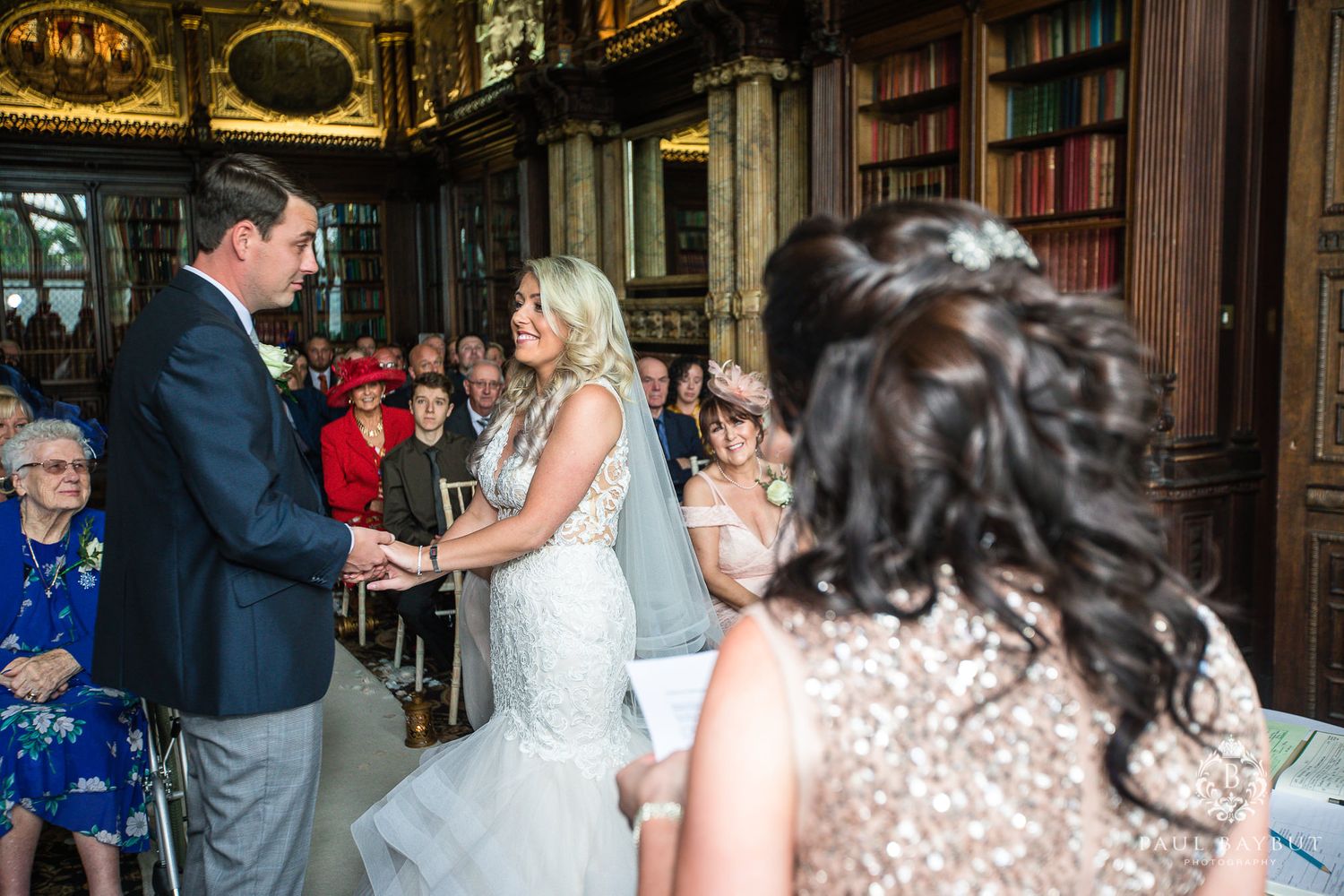 Couple exchanging rings during intimate library wedding with guests looking on.