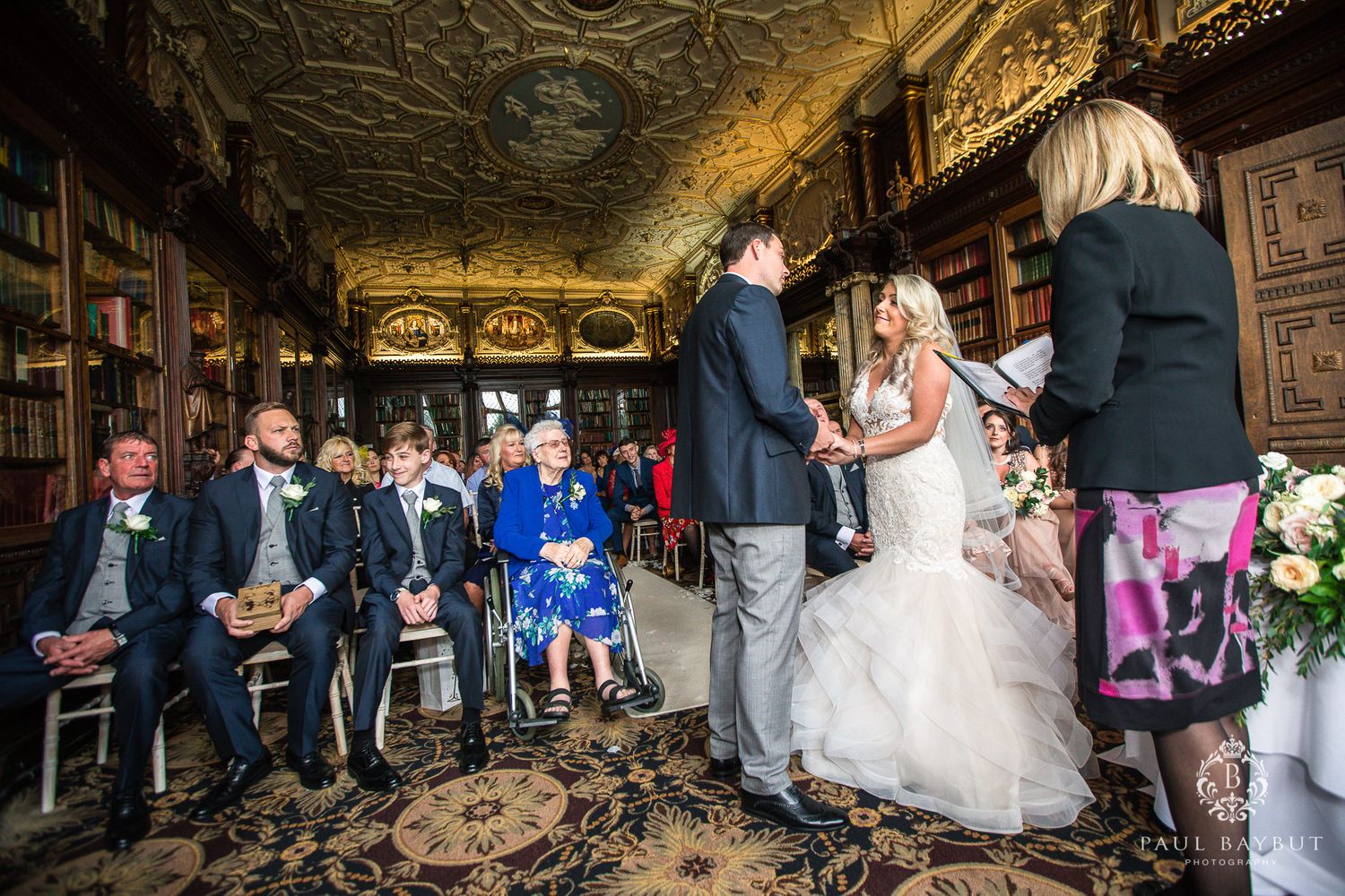 Wide view of a wedding ceremony in a grand historic library with ornate ceiling and decor.