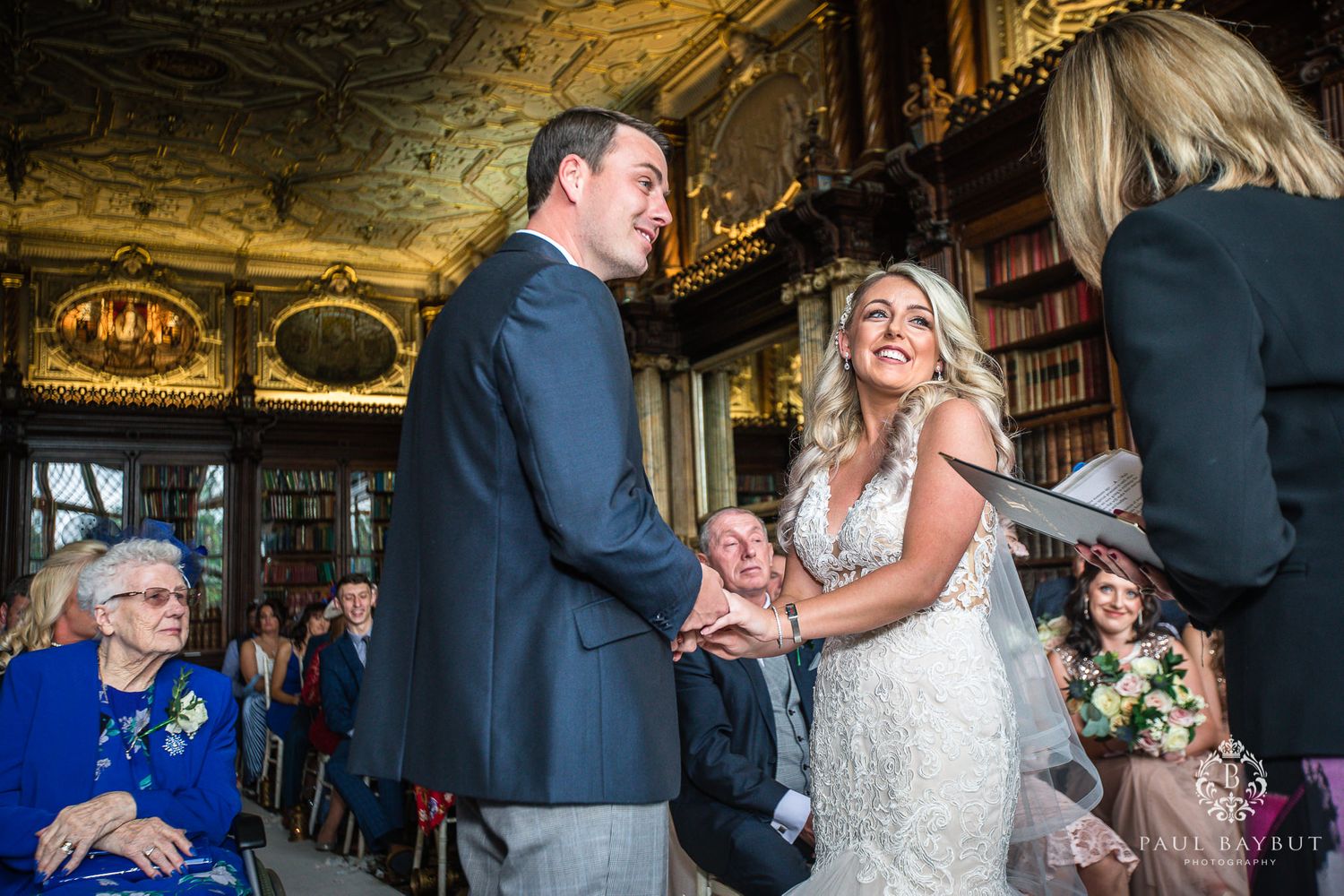 A wedding ceremony takes place in an ornate library with bookshelves and chandeliers.
