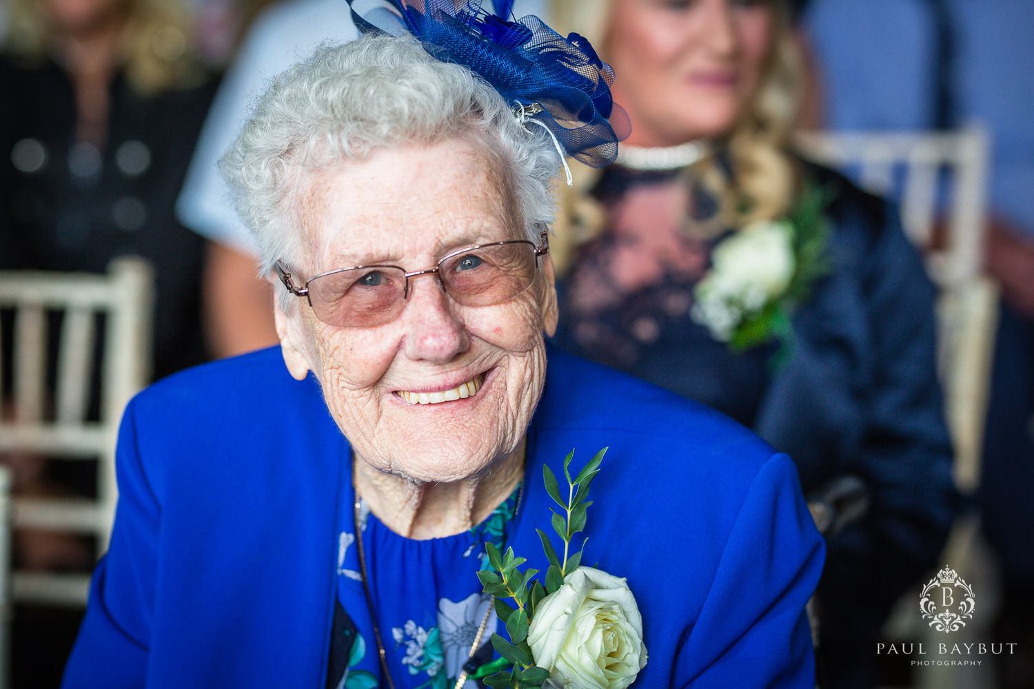 Senior guest in bright blue attire smiles while wearing a matching fascinator at formal event.