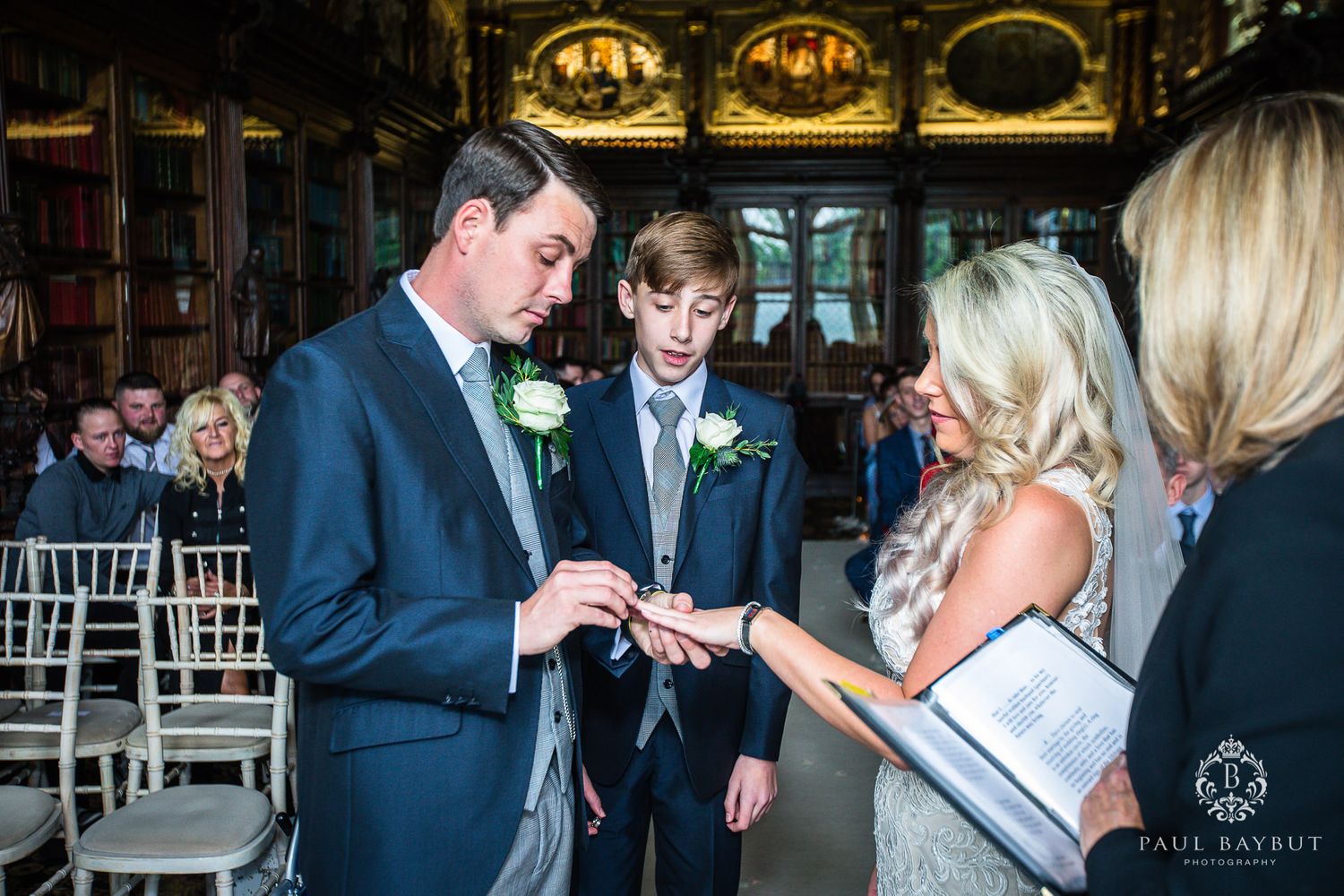 Ring exchange during wedding ceremony in grand library with ornate decor.