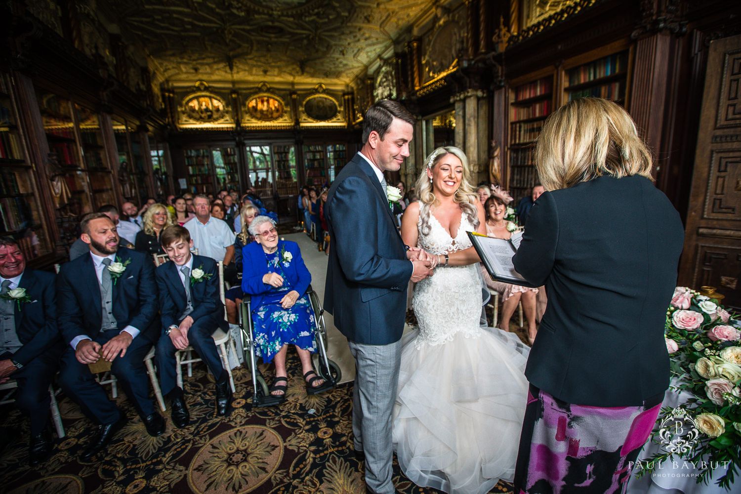 Wide angle view of wedding ceremony in historic library with gathered guests.