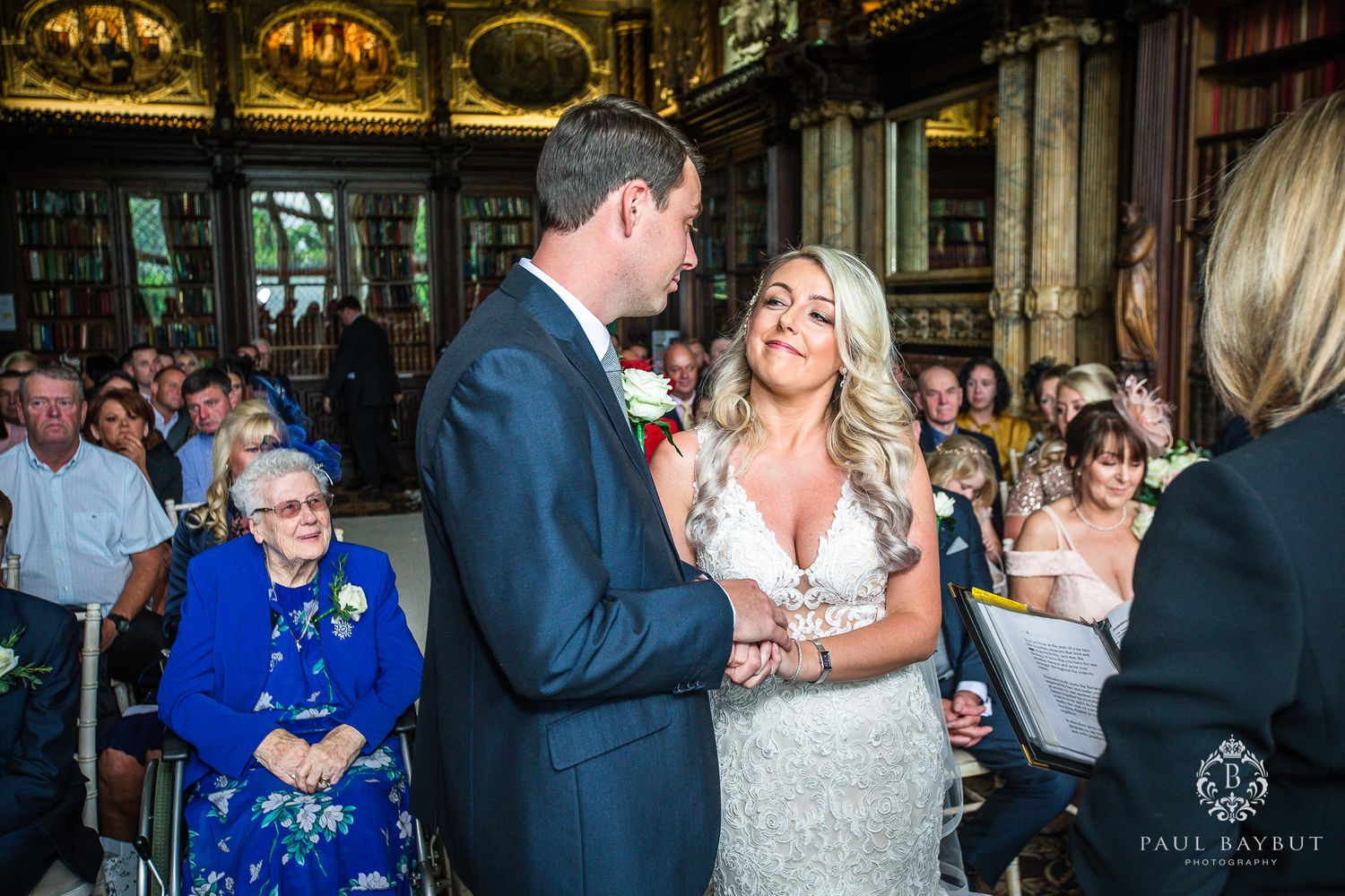 A joyous wedding ceremony unfolds in an ornate library with elegant decor and guests looking on.