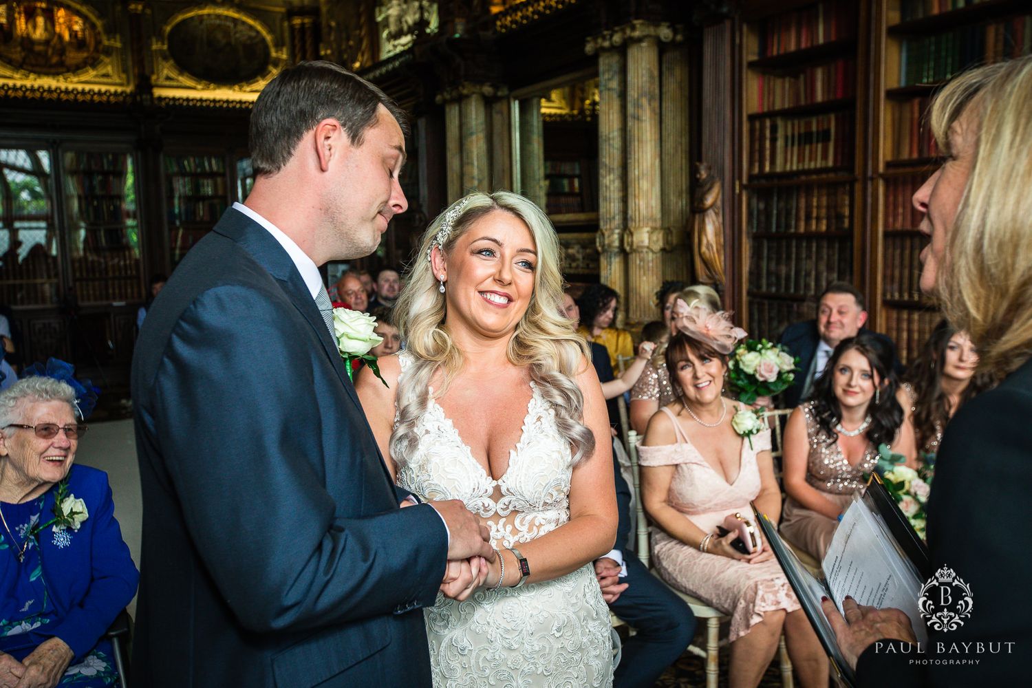 Wedding vows being exchanged in an elegant library with guests watching from wooden chairs.