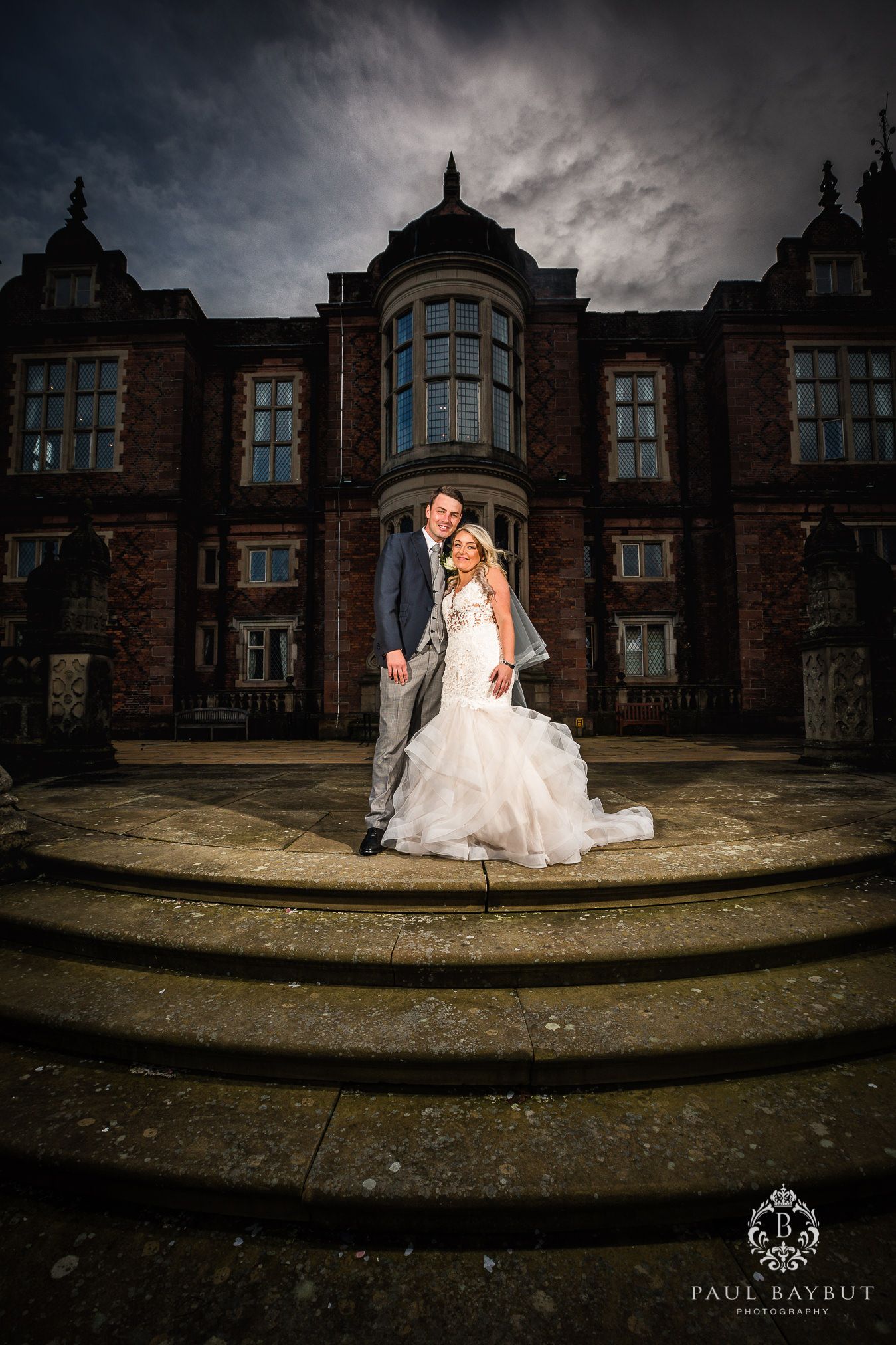 A dramatic wedding photo against a historic brick mansion with curved steps under stormy skies.