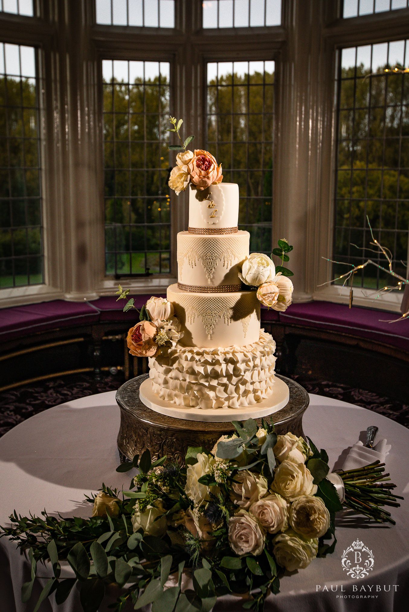 An elegant four-tier white wedding cake decorated with roses displayed in a bay window setting.