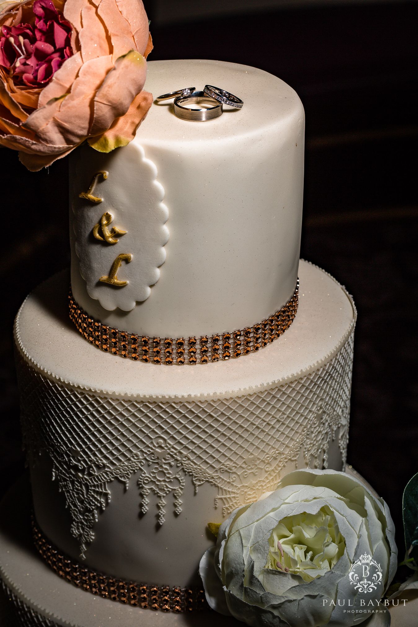 Elegant three-tier white wedding cake with metallic accents, floral decorations, and wedding rings on top.